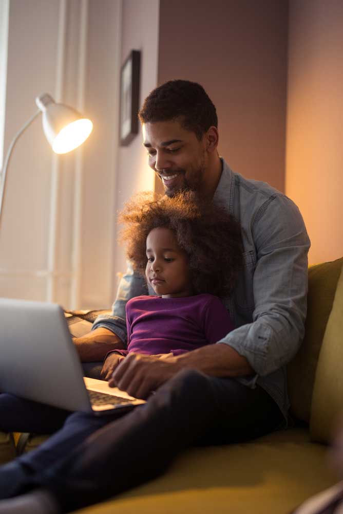 Smiling Father Sitting With Daughter On A Chair Working On Laptop - Contact Us To Learn More About Under 65 Health Insurance Services By Agape Insurance And Financial Group In Tupelo, Mississippi